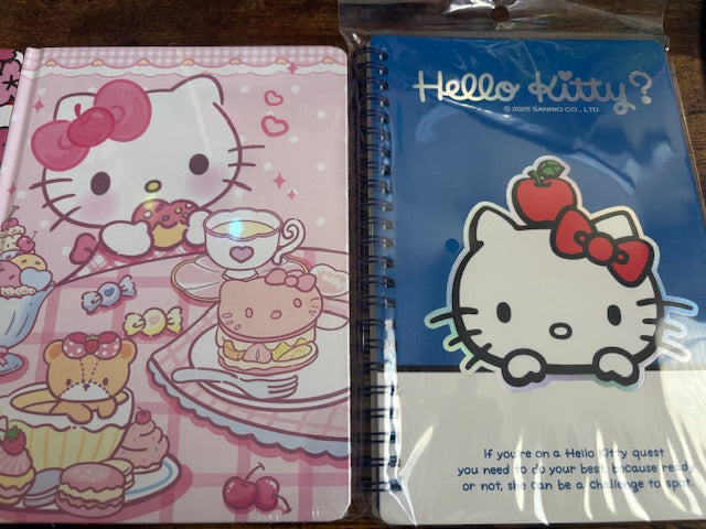 Two Hello Kitty notebooks, one pink with cartoon characters and the other blue with a face design, on a wooden surface.
